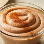 The image shows a delicious bowl of chocolate pudding. The pudding has a smooth and creamy texture with a rich chocolate color. It is beautifully swirled on top, giving it an elegant, homemade look. A light dusting of cocoa powder or cinnamon adds a finishing touch, making the dessert even more visually appealing. The pudding is served in a clear glass bowl, with a spoon resting beside it, ready to dig in. This dessert looks indulgent, perfect for a comforting treat after a meal or as a special dessert.